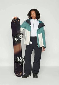 Snowboardeur portant une veste imperméable teal et blanche, un pantalon noir et des bottes, tenant un snowboard noir avec des fixations blanches.