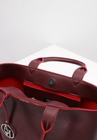 Vínová kabelka s texturovaným povrchem, kontrastním červeným vnitřkem, dvěma držadly a zapínáním na patent. Obsahuje logo a vnit�řní kapsu.