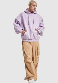 Lilla oversized hettegenser med frontlomme og hvit logo, kombinert med brune cargo bukser med flere lommer og en avslappet passform.