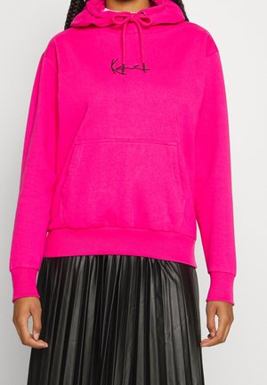 Persoon draagt een felroze hoodie met voorzak en een zwarte geplooide rok, staand tegen een effen lichte achtergrond.