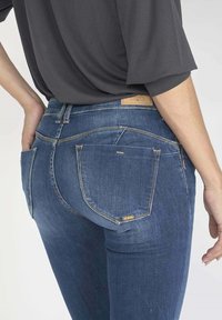 Jean en denim bleu foncé avec une coupe slim, présentant des poches arrière avec des coutures visibles et une étiquette en cuir marron clair sur la taille.