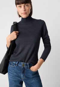 Mørkeblå turtleneck laget av et mykt materiale, kombinert med høy midje blå jeans. Modellen holder en svart skulderveske.