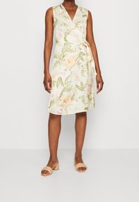 Bloemen wikkeljurk in beige met groene bladeren en pastelachtige bloemen. Mouwloos ontwerp, V-hals en strikceintuur. Gekomplimenteerd met lichtgekleurde sandalen.