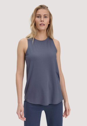 Blonde vrouw die een mouwloos, donkergrijs sporttopje en marineblauwe leggings draagt, staand tegen een effen lichte achtergrond.