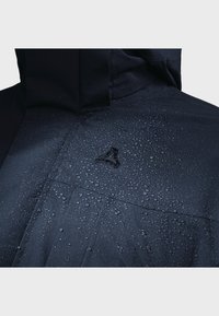 Marineblauer wasserdichter Mantel mit strukturiertem Oberfläche, auf dem Wassertropfen sichtbar sind, und einem markanten Logo in der Mitte. Robustes Material und Designdetails sind erkennbar.