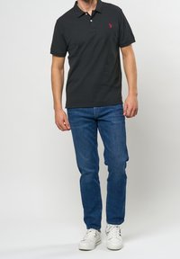 Mörkgrå polo t-shirt med korta ärmar, tvåknapprig krage och röd logotyp, parat med blå jeans och vita sneakers.