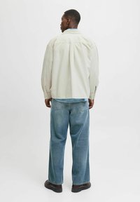 Mand set fra baggrunden iført cremefarvet jakke, blå denimskjorte, løse blå jeans og mørkebrune sko, stående på en lys hvid baggrund.