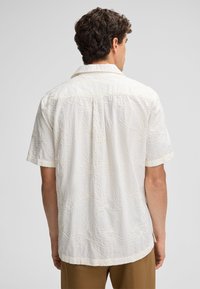 Korte mouwen, crèmekleurig shirt met een textuur van bloemachtige borduursels, knoopsluiting met kraag en een rechte zoom. Natuurlijke stofafwerking.