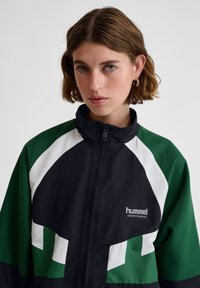 Young woman with short brown hair wearing a green, black, and white Hummel sportswear jacket with a front zipper, facing forward against a plain background.