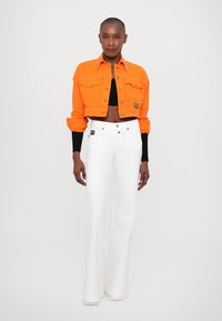 Giacca arancione a trama cropped con bottoni frontali e due tasche sul petto, abbinata a pantaloni bianchi a gamba larga con bottoni in tonalità oro.