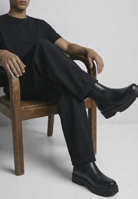 Stivaletti alla caviglia in pelle nera con suola spessa, indossati con pantaloni neri e una t-shirt nera, seduti su una sedia di legno. Texture liscia.