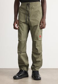 Mann in olivgrünen Cargo-Hosen mit elastischem Bund und schwarzem Kordelzug, kombiniert mit glänzenden schwarzen Sneakers, steht auf grauem Boden.