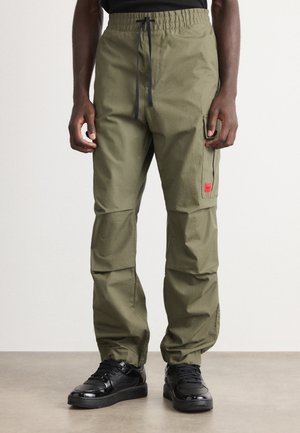Man wearing olive green cargo pants with elastic waist and black drawstring, paired with black shiny sneakers, standing on gray floor.