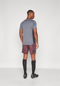 Idrottskläder med en grå kortärmad tröja, bordeauxfärgade mönstrade shorts, svarta knästrumpor och svarta fotbollsskor.
