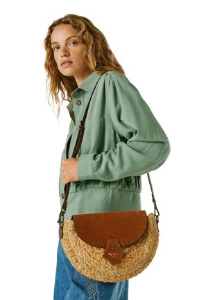 Vrouw met golvend haar draagt een groene jas met knopen en blauwe spijkerbroek, draagt een geweven ronde crossbodytas met een bruine leren klep.