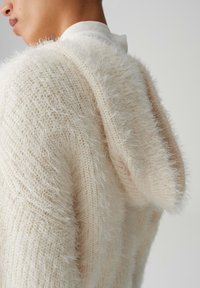 Cremefarbener Kapuzenpullover mit weicher, flauschiger Textur und lockerem Strickdesign, ausgestattet mit einer markanten Kapuze und dezenten Musterdetails.