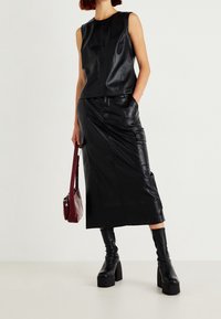 Schwarzes ärmelloses Lederoberteil, kombiniert mit einem langen Lederrock. Schwarze knielange Stiefel und eine kleine burgunderfarbene Handtasche vervollständigen das Outfit.