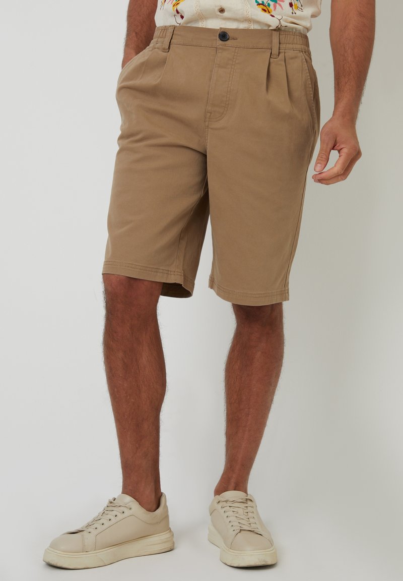 Khaki-Shorts mit einem elastischen Bund, gefaltetem Frontbereich, seitlichen Taschen und geradem Schnitt; kombiniert mit hellen Sneakers.