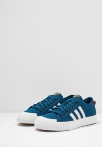 Pár modrých nízkých tenisek Adidas s bílou podrážkou, bílými pruhy s červeným prošíváním a žlutými štítky s logem na jazyku a červenými patními poutky.