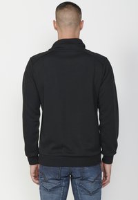 Suéter negro texturizado con cuello alto y sutil patrón de espiga, complementado con puños y dobladillo acanalados.