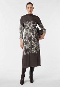 Braunes Schlangenprint-Midikleid mit langen Ärmeln und hohem Kragen. Glatter Stoff, tailliertes Oberteil und ausgestellter Rock. Das Model hält eine schwarze Tasche.