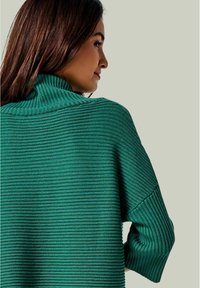 Grüner gerippter Pullover mit hohem Kragen, ausgestattet mit horizontalen Streifen und locker sitzenden Ärmeln, präsentiert vor einem neutralen Hintergrund.