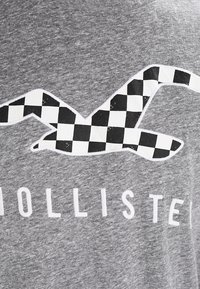 Šedé tričko s černobílým kostkovaným designem racka. Materiál vypadá měkce, má klasický kulatý výstřih a potisklý bílý text níže.