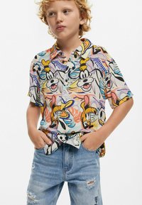 Camicetta colorata a maniche corte con personaggi dei cartoni animati, tra cui Paperino, su uno sfondo di un motivo tropicale. Il tessuto sembra leggero; abbinata a jeans strappati.