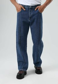 Jean en denim bleu avec une coupe droite, coutures visibles le long des coutures latérales et design à cinq poches. Porté avec des chaussures noires.