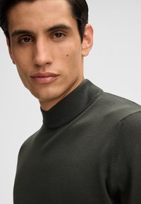 Dunkelgrüner, gerippter Rollkragenpullover mit tailliertem Design. Glatte Textur mit hohem Kragen. Nahaufnahme des Ausschnitts und des Gesichts.
