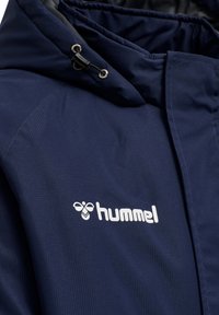 Marinblå jacka i texturerat tyg; har en huva, dragkedja och en vit logotyp på bröstet. Accenter inkluderar justerbara snoddar.