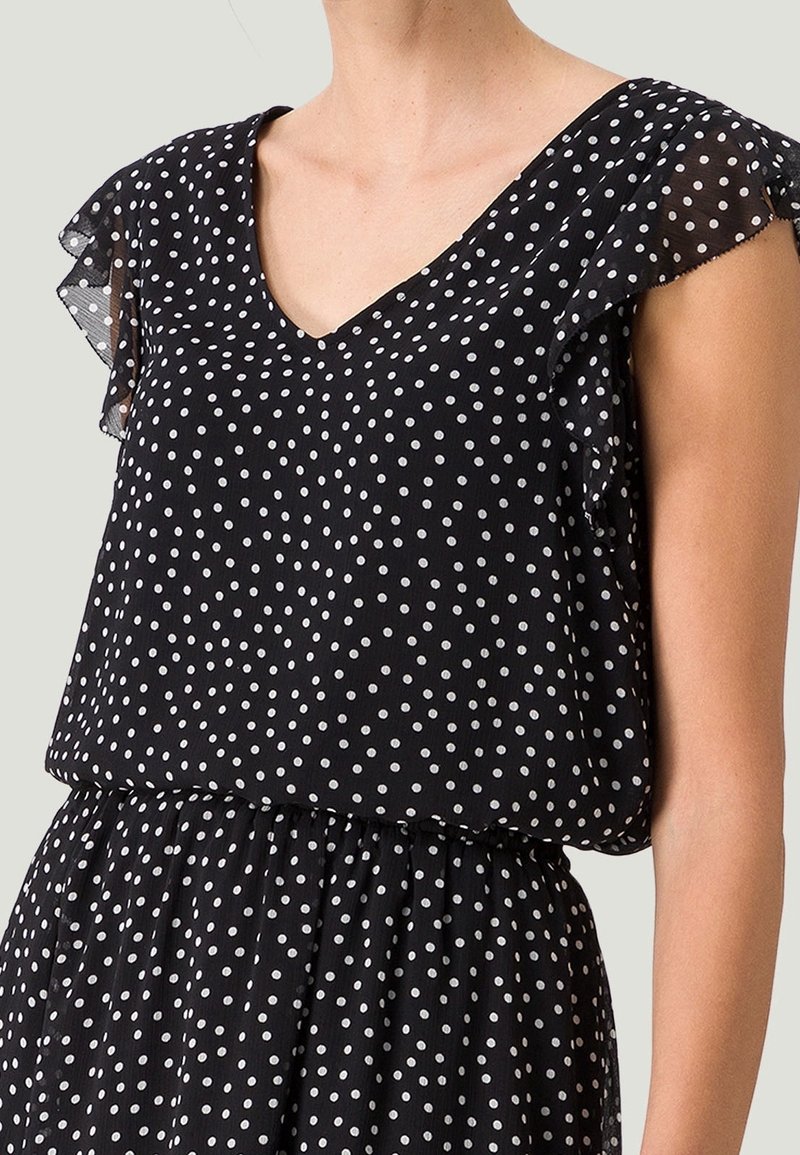 Robe noire sans manches avec des pois blancs et des détails volantés aux épaules sur une personne, du cou à la taille.