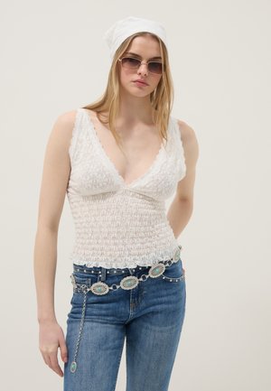 Jeune femme portant un haut sans manches en dentelle blanche, un jean bleu avec une ceinture argentée décorative, un foulard blanc sur la tête et des lunettes de soleil.