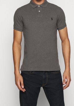 Torse masculin portant un polo gris foncé à manches courtes ajusté et un jean noir, debout avec les mains détendues le long du corps.