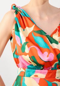 Femme portant une robe sans manches avec un imprimé floral abstrait vif en orange, sarcelle, rose, vert et crème, détail du nœud à l'épaule visible.