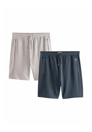 Młody mężczyzna stoi z rękami w kieszeniach, ubrany w białą koszulkę, granatowe spodenki, białe trampki, skarpetki i szarą bluzę zarzuconą na ramiona.