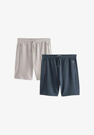 Junger Mann steht mit den Händen in den Taschen, trägt ein weißes T-Shirt, marineblaue Shorts, weiße Turnschuhe, Socken und einen grauen Pullover, der über die Schultern gelegt ist.