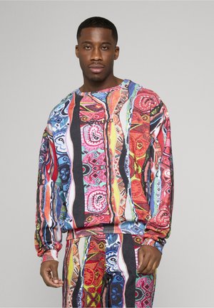 Mand iført farverig, mønstret, matchende sweatshirt og bukser med abstrakte designs, stående mod en ensartet lys grå baggrund.