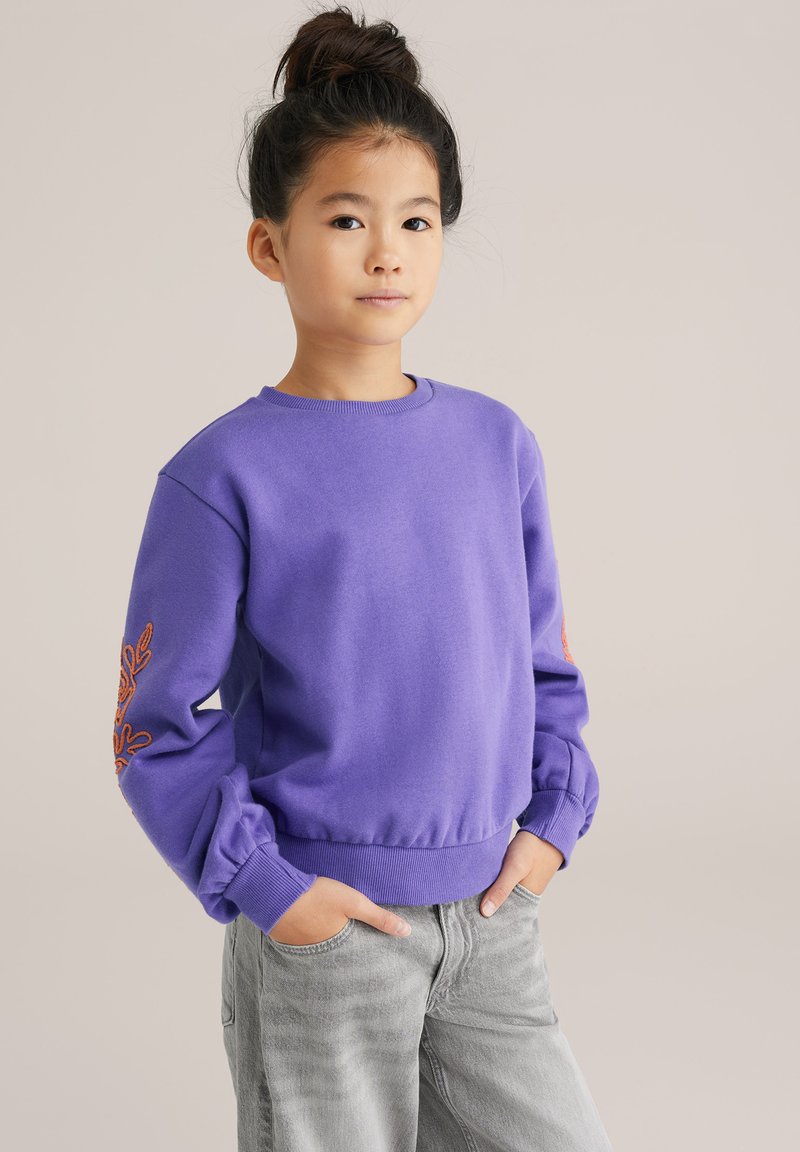 Pulover violet cu mâneci lungi și puf, având manșete strâmte, cu broderie florală portocalie pe mâneca stângă. Purtați cu blugi gri.