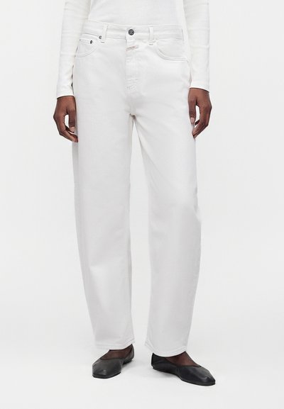 Personne portant un jean blanc droit, des chaussures plates noires et un haut blanc à manches longues, debout devant un fond blanc uni.
