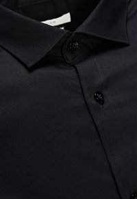 Svart skjorta med knappar i mjukt tyg som har en skarp krage, två framtill placerade knappar och diskreta sömnadsdetaljer.