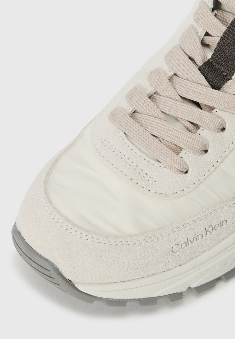 Zapatillas de color claro que combinan suave tejido y texturas de ante. Los cordones son de un tono más claro, con un logo sutil en el lateral.