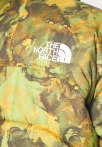 Veste camouflage verte et marron avec le logo blanc The North Face sur la partie supérieure du dos.