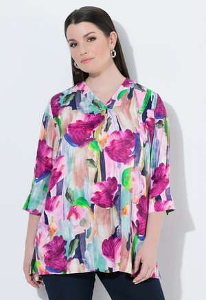 Kvinde med lige mørkt hår iført sølvøreringe i ringform og en farverig bluse med blomster i pink, grønne og blå toner, der står op ad en hvid væg.