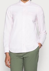 Camicia a maniche lunghe rosa chiaro con sottili righe bianche, colletto button-down, taschino sul petto e orlo dritto. Abbinata a pantaloni verdi.