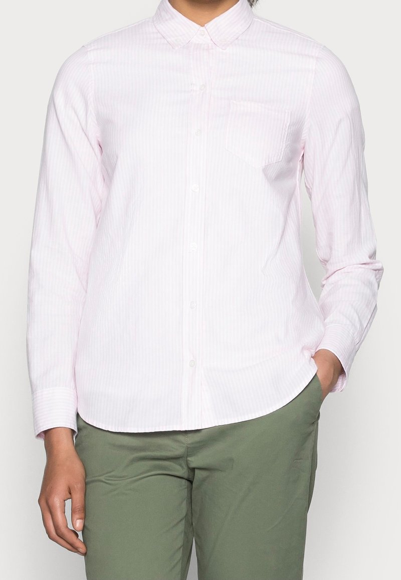 Camicia a maniche lunghe rosa chiaro con sottili righe bianche, colletto button-down, taschino sul petto e orlo dritto. Abbinata a pantaloni verdi.