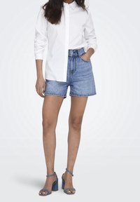 Eine Frau trägt ein weißes Hemd mit Knöpfen, hochgeschnittene blaue Jeansshorts und blaue Absatzsandalen. Sie steht mit einer Hand in der Tasche.