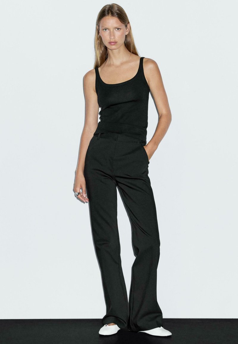 Czarny top na ramiączkach i dopasowane czarne spodnie, wyróżniające się dopasowanym fasonem i minimalistycznym designem. Ubrane z białymi butami typu slip-on, stojące na czarnym tle.