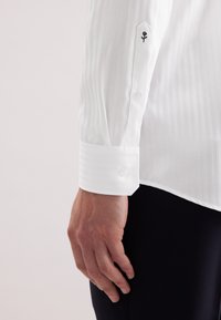 Camicia bianca con sottili righe verticali, dotata di polsini con bottoni e un dettaglio di rosa nera ricamata sulla manica.