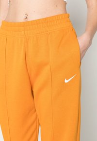 Orange sweatpants med elastisk midja och sidofickor; har en vit Nike-logga på vänster lår. Mjuk tyg och avslappnad passform.